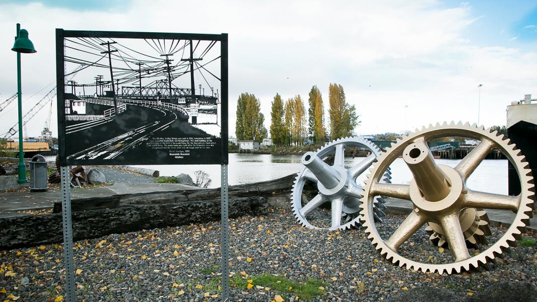 Outdoor sculpture installation with a metal sign and large gear sculptures near the water.