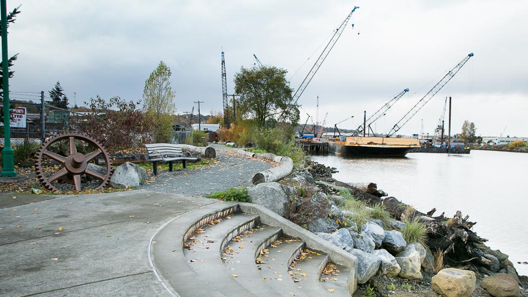 Waterfront path with benches, large metal gear sculpture, and cranes across the water.