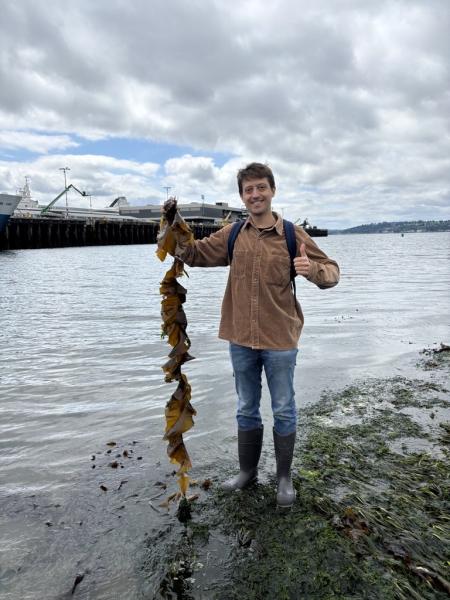 Jack stands on a shoreline holding a long strand of seaweed, with water and a docked vessel in the background.