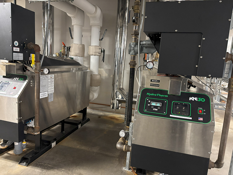 Industrial equipment, including two stainless‑steel heating or boiler units with connected pipes and insulated lines, sits in a mechanical room with concrete floors and exposed infrastructure.