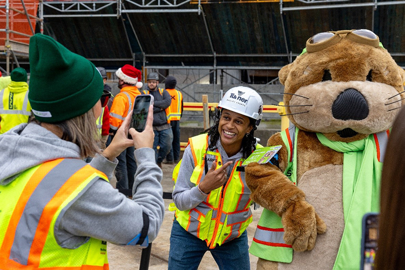 A person in construction safety gear poses for a photo with JETT the otter mascot at an active jobsite while another person takes the picture.