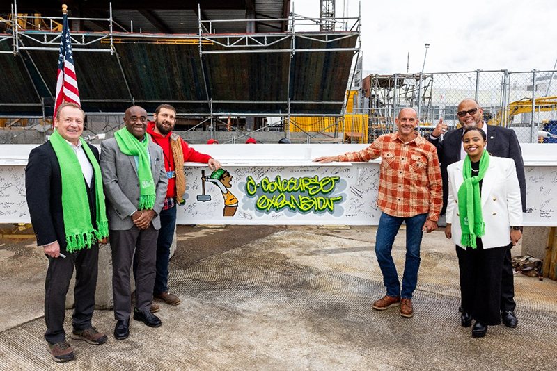Six people stand in front of a signed construction beam painted with “C Concourse Expansion,” positioned at an active jobsite with equipment and scaffolding in the background.