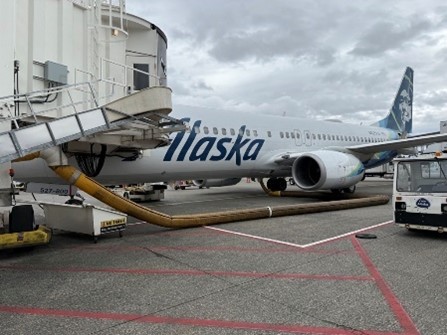 An Alaska‑branded airplane is parked at an airport gate, with a jet bridge connected to the aircraft and service equipment positioned nearby on the tarmac.