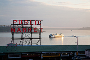 A large red Public Market sign overlooking a calm waterfront at sunset, with a ferry crossing in the background.