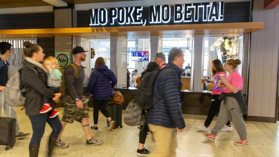 Travelers walk past the Poke to the Max restaurant at SEA Airport, with the sign “MO POKÉ, MO BETTA!” displayed above the storefront.
