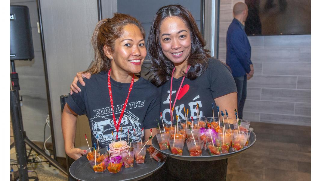 Two individuals stand indoors holding trays of small poke samples arranged in clear cups with garnish, offering food during the Poke to the Max opening event at SEA Airport.