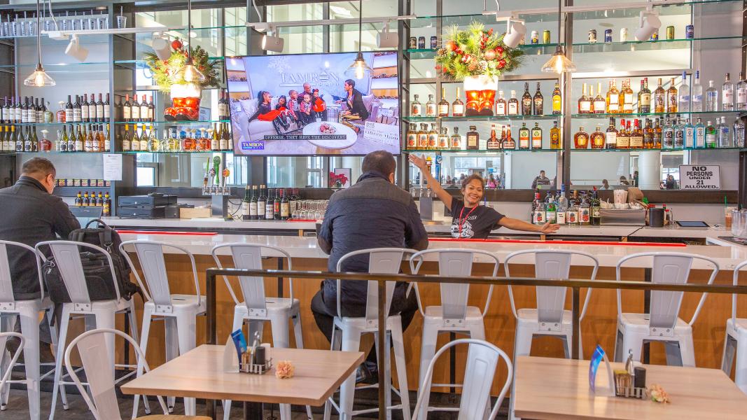 A bar area inside Poke to the Max at SEA Airport with rows of bottles displayed on shelves, a large television mounted above the counter, and seated customers. A staff member behind the bar extends an arm outward while interacting with guests.
