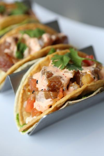 A close-up of a poke taco in a metal holder, filled with diced marinated fish, sauce, cilantro, and small vegetable pieces, with additional tacos visible behind it.