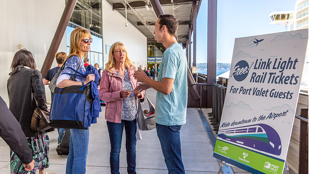 "People stand outside a building near a large sign that promotes free Link light rail tickets for Port Valet guests. One person is speaking with two others who are holding bags. Additional travelers are visible in the background, and the walkway overlooks the water.