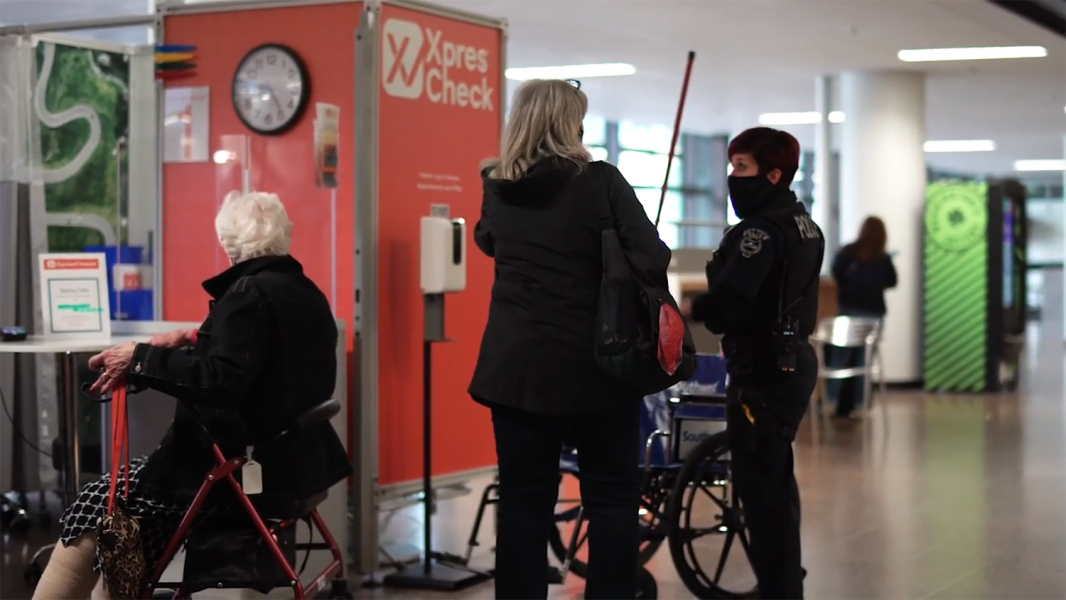 A security officer speaking with two travelers near an XpresCheck station, including one person seated with a walker.