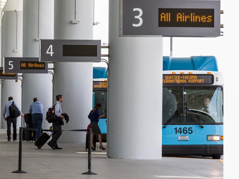 Shuttle buses parked at numbered pick-up zones outside a terminal, with several travelers walking past the tall white columns and digital signs displaying “All Airlines.