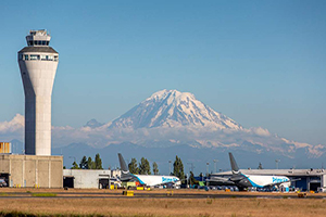 Airport airfield with a control tower, parked airplanes, and a snow-covered mountain in the background.