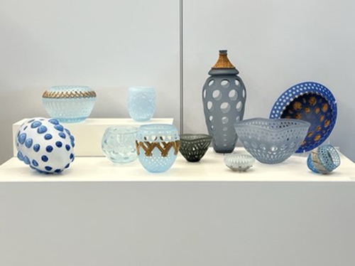 A group of blue, white, and gray glass vessels with dotted and cutout patterns displayed on a white pedestal.