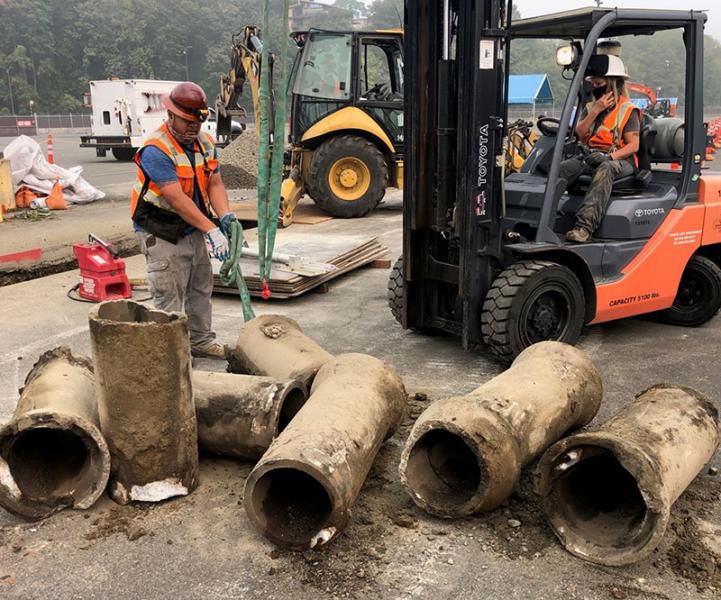 Workers use a forklift and straps to move several old concrete pipes at a construction site with heavy equipment in the background.