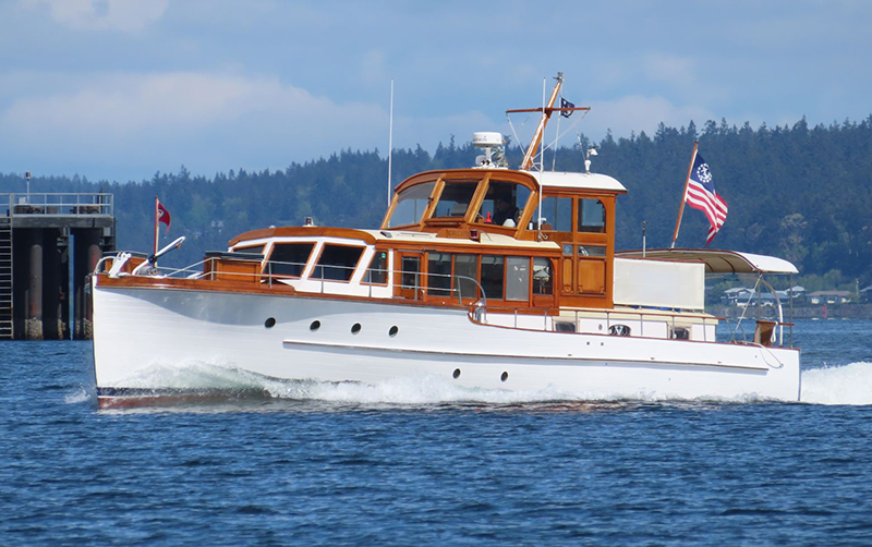 A white boat named “Riptide” moves across the water, featuring wood‑trimmed cabin structures, multiple windows along the hull, and an American flag at the stern, with a tree‑covered shoreline and cloudy sky in the background.
