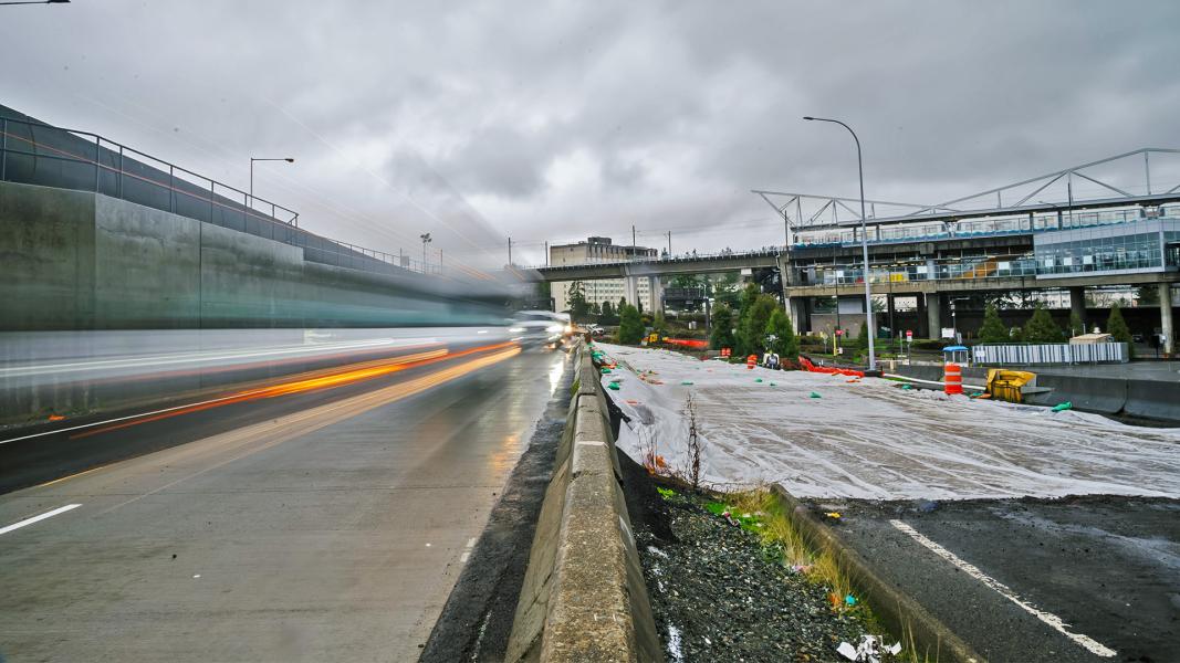 Blurred headlights streak along a wet road at SEA Airport beside a construction area covered with tarps and orange barrels, with an elevated walkway and buildings in the background under cloudy skies.