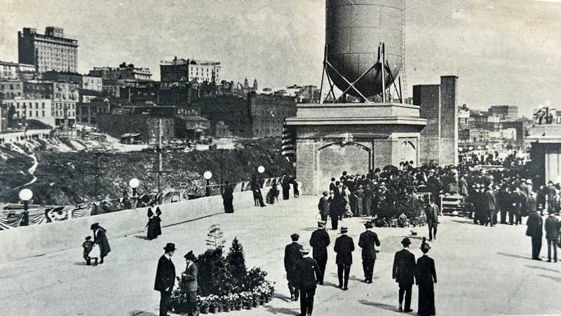 A historic photograph of people walking and gathering on a waterfront promenade with city buildings on the hillside behind them.