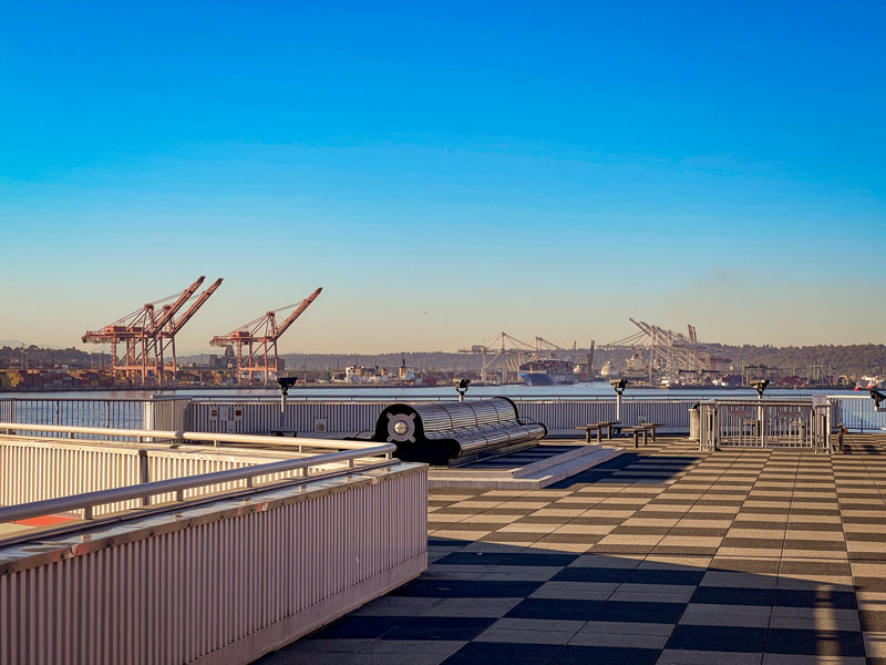 A waterfront deck with benches and railings overlooks a harbor with large cargo cranes in the distance under a clear blue sky.