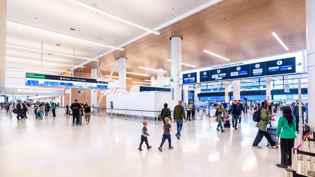 Travelers walk through a bright, spacious SEA Airport concourse near security checkpoints, with overhead signs directing people to gates and screening lanes.