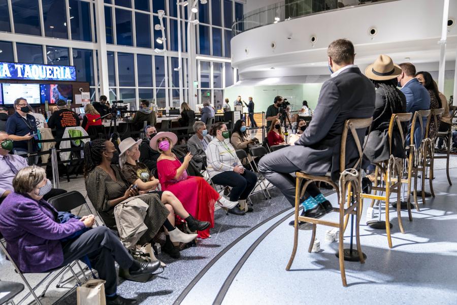 Audience seated indoors watching a masked panel speak at the front in a bright, modern event space.