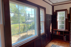 Interior room with large, wood‑trimmed windows showing a bright outdoor view of trees, grass, and a hedge. The room has dark wood paneling, built‑in shelving with a few books, and sunlight coming through the windows onto the hardwood floor.