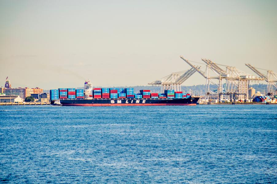 The cargo ship SM Seoul moves across a harbor carrying stacked red and blue containers. Large loading cranes line the port in the background, with a cityscape visible under clear skies.