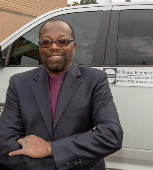 Samuel Obunike wearing a dark blazer and glasses stands with arms crossed in front of a white vehicle displaying a company sign that reads “O’Bunco Engineering” along with contact information.