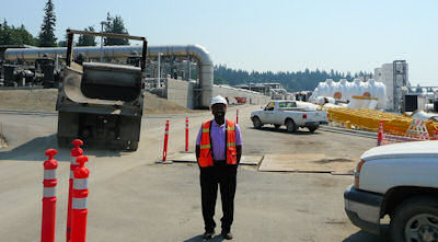 Samuel wearing a reflective safety vest and a hard hat stands on a roadway at an industrial construction site with heavy equipment, vehicles, and piping in the background.