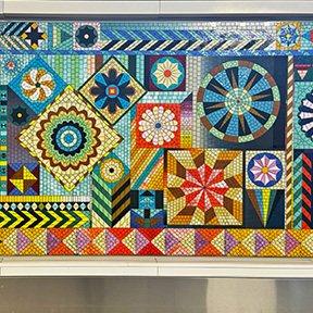 The artwork “On a Clear Day You Can See Forever” by Sarah More is a brightly colored mosaic-style piece made up of geometric shapes and patterns, including circles, diamonds, zigzags, and star-like designs in vibrant blues, greens, yellows, reds, and oranges.