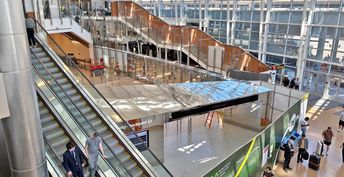 Airport terminal view with escalators, upper walkways, and a security checkpoint area under construction.