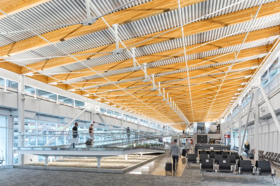 Interior of an airport concourse with a high ceiling made of wood and metal beams, a glass walkway with travelers moving across it, and seating areas below.