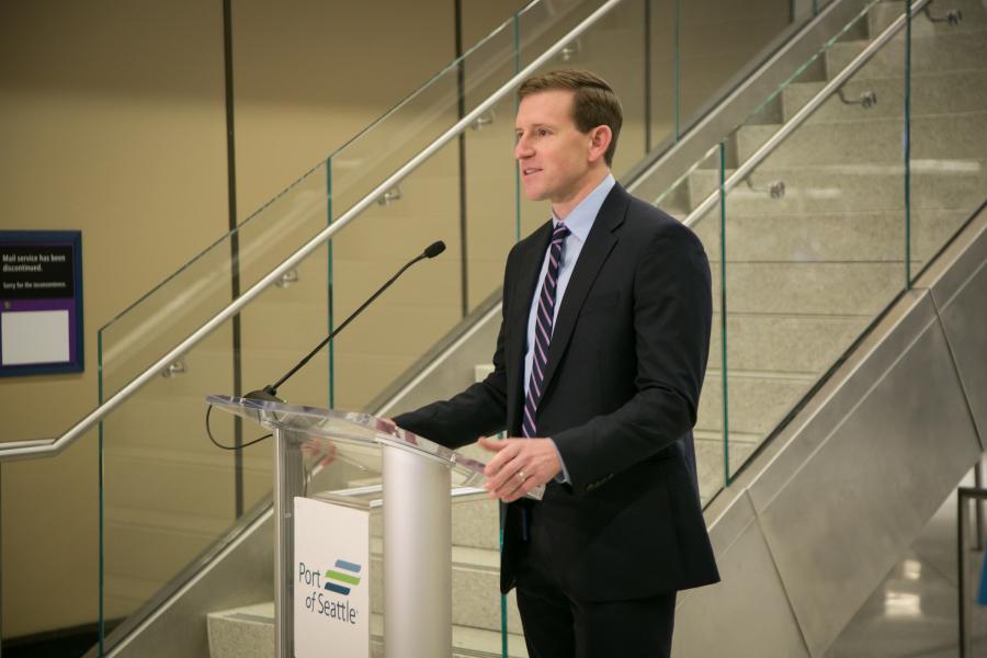 Commissioner Ryan Calkins speaking at a clear podium with a microphone inside a modern building, with glass‑railed stairs and signage visible in the background.