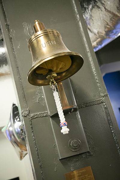 A brass bell marked “USO” hangs on a metal post, with a braided white pull cord attached beneath it. The bell is mounted indoors with nearby framed items and metal ductwork visible.