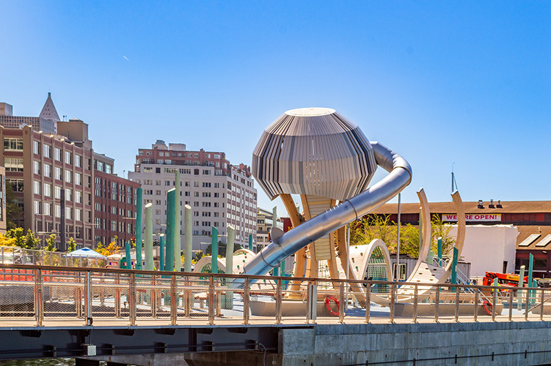 Playground at Waterfront Park featuring a tall dome-like climbing structure with a long metal slide, set against nearby buildings and a clear blue sky.