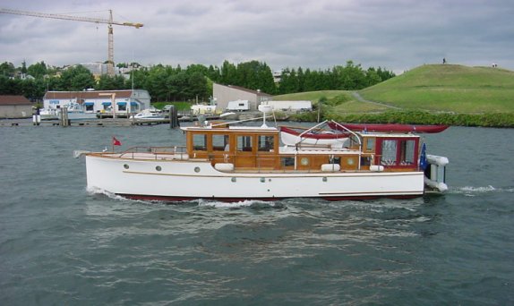 A white boat named “Seven Bells” moves across the water, featuring wood‑trimmed cabin structures and an open deck area, with a shoreline containing buildings, a grassy hill, and a construction crane in the background under a cloudy sky.