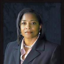 Sharon Black in a dark blazer and light shirt sits against a dark background, looking toward the camera.