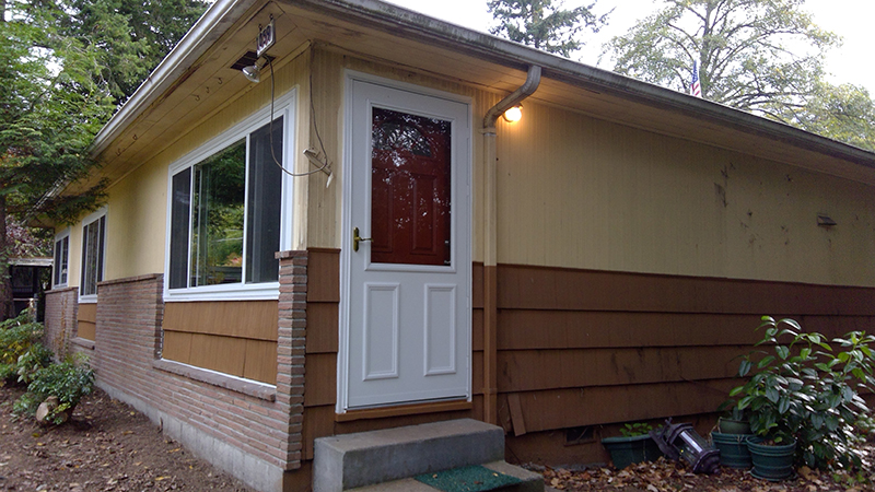 Exterior view of a single‑story house with light-colored siding on top and brown siding below, featuring a white door with a small concrete step and windows along the side. The house is surrounded by trees and plants.
