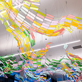 The artwork “Rain Shadow” by Soo Sunny Park consists of suspended, flowing panels made of translucent, colorful materials in shades of pink, yellow, green, and blue. The pieces form wavy, ribbon-like shapes that hang from the ceiling, creating a layered, shimmering installation.