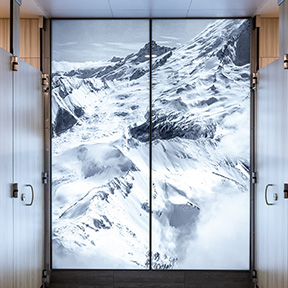 A large wall-mounted photographic artwork by Spike Mafford and Nikita Mehta shows a detailed, high‑contrast view of snowy mountain peaks and deep ridges. The image is displayed on two tall panels positioned between smooth metal and wood surfaces in an indoor hallway.
