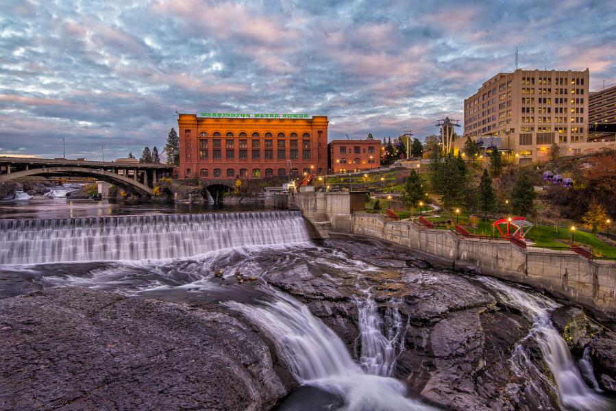 A river flows over a series of falls in a downtown area, with a large brick building and bridges under a cloudy sky.