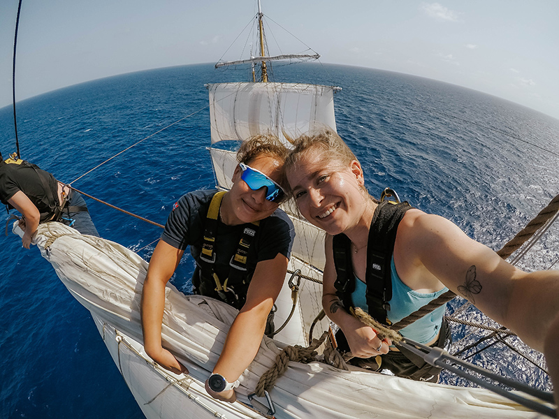 Two people wearing safety harnesses sit on a large sail high above the ocean aboard the tall ship Statsraad Lehmkuhl. They are positioned near the rigging with the open sea in the background, and one person holds the camera for the photo.