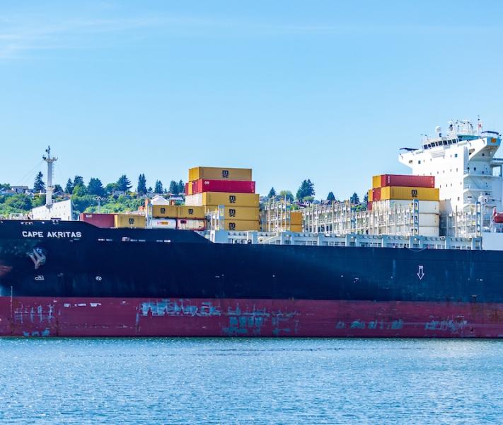 The container ship Cape Akritas is docked at a port, carrying stacked yellow and red cargo containers.