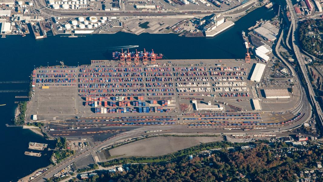 Aerial view of a large shipping terminal with rows of stacked cargo containers, multiple container cranes along the waterfront, adjacent industrial buildings, and rail lines running along the lower edge of the site.