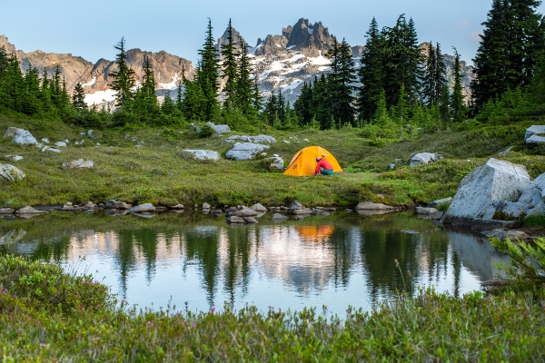 An orange tent set beside a small reflective pond in an alpine meadow, surrounded by evergreen trees and snow‑dusted mountains.