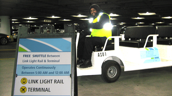 A person wearing a reflective safety vest sits in a white airport shuttle cart parked indoors next to a sign. The sign reads “FREE SHUTTLE Between Link Light Rail & Terminal,” with details stating it operates continuously between 5:00 AM and 12:00 AM, and includes icons for Link Light Rail and Terminal.