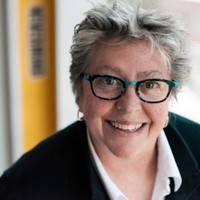 A close-up headshot of Trish Drew indoors, wearing glasses and looking toward the camera.
