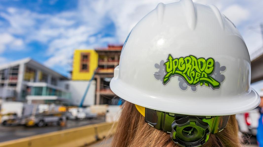 A person wearing a white hard hat labeled “Upgrade SEA” stands at a SEA Airport construction site, with partially built structures and equipment visible in the background.