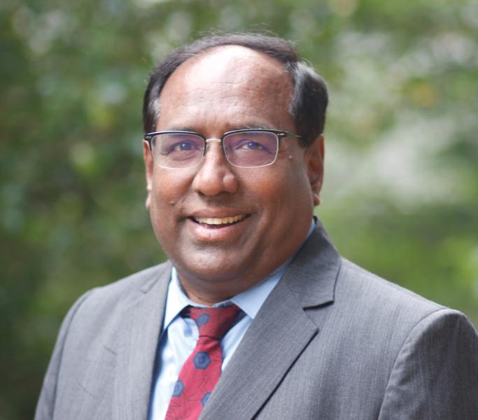 Venky Ramakrishnan wearing glasses, a suit jacket, a collared shirt, and a patterned tie stands outdoors with blurred greenery in the background, looking toward the camera.