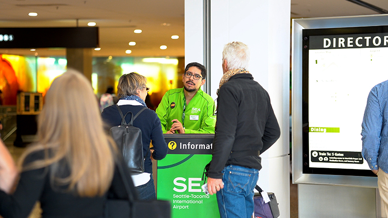 Airport information desk volunteer in a green SEA Seattle‑Tacoma International Airport shirt assists travelers beside a terminal directory sign.