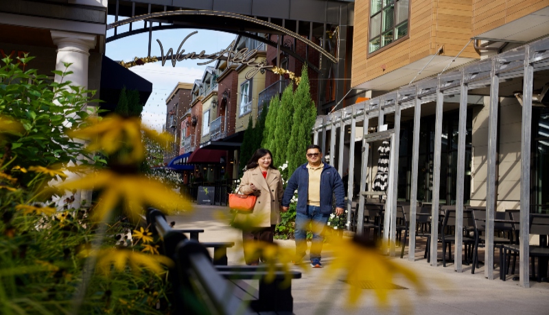 Two people walk through Woodinville’s Wine Alley, with yellow flowers in the foreground and storefronts lining the outdoor pathway.